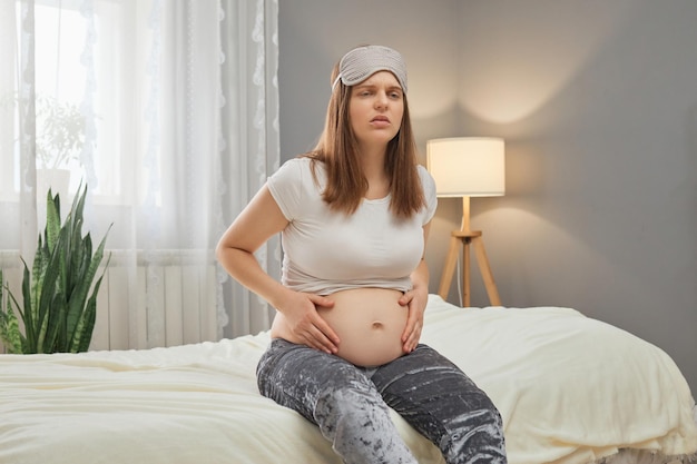 Foto ongezonde zwangere vrouw die haar buik vasthoudt terwijl ze thuis op bed zit, lijdt aan spasmen, begint met samentrekkingen, grimast van pijn, draagt een pyjama en een slaapmasker.