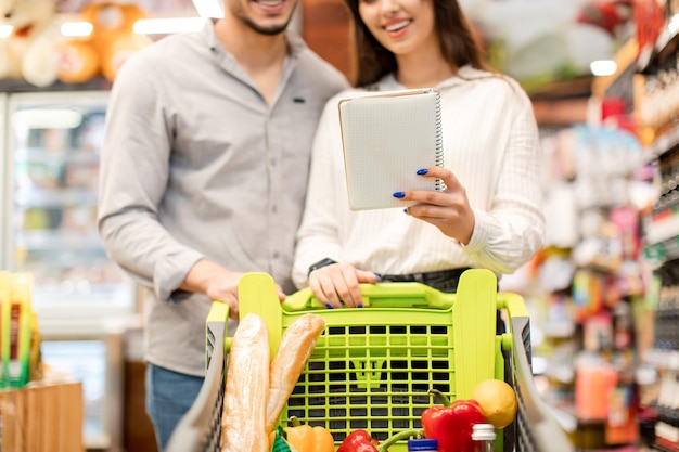 Onherkenbaar paar dat een boodschappenlijstje vasthoudt om voedsel in de supermarkt te kopen