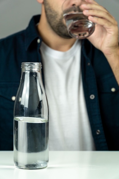 Onherkenbare bebaarde man drinkwater uit een glazen tuimelaar met een halfvolle glazen fles op de voorgrond gezondheid en hydratatie concept