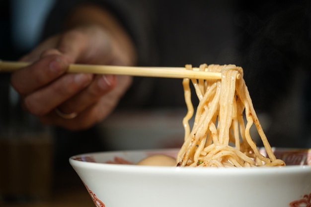 Onherkenbare gewaspersoon die eetstokjes gebruikt tijdens het eten van smakelijke ramen met noedels uit een witte kom op onscherpe achtergrond