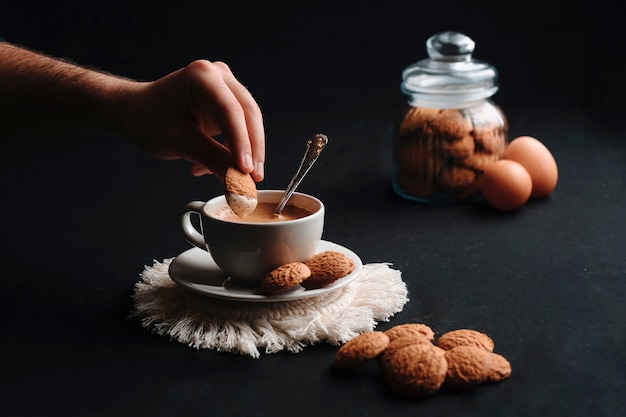 Onherkenbare hand die een koekje onderdompelt in hete koffie met koekjes in een kruik en eieren. Detailopname.