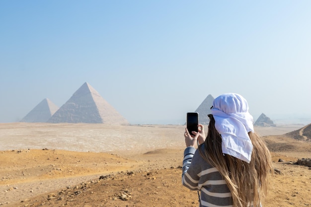 Onherkenbare jonge toeristenvrouw van achteren met witte tulband op het hoofd die een foto maakt van de piramides van Caire, Egypte