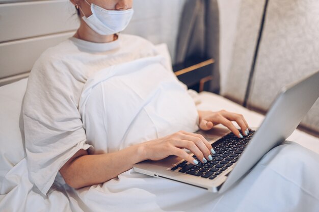 Onherkenbare jonge vrouw in het masker van de gezichtsgeneeskunde die aan laptop in slaapkamer werken tijdens de quarantaine van het coronavirusisolatiehuis. Covid-19 pandemisch Corona-virus. Afstand online werk vanuit huis concept.