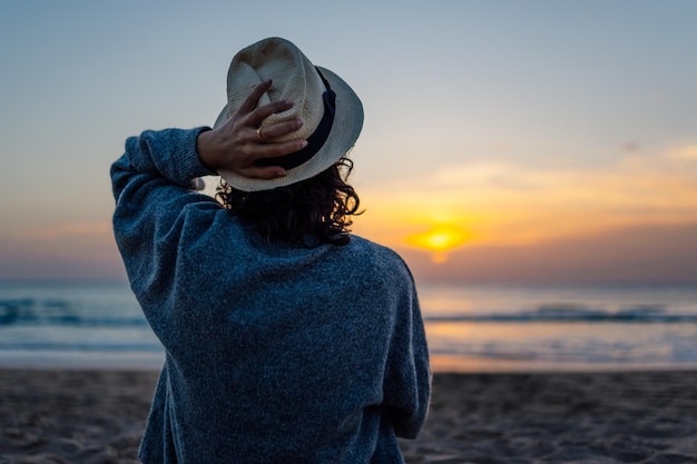 Onherkenbare jonge vrouw in hoed die van zonsondergang op het strand geniet