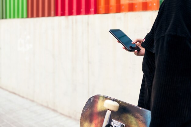 Onherkenbare jonge vrouw typt op de mobiele telefoon terwijl ze over straat loopt met haar skateboard in haar hand