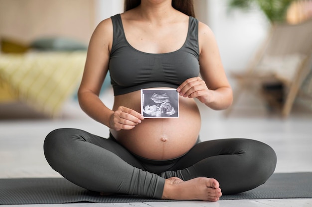 Foto onherkenbare jonge zwangere dame die op yogamat zit en babysonografie demonstreert