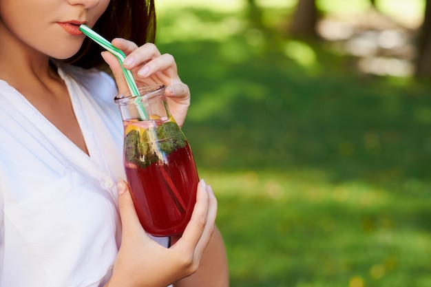 Onherkenbare jongedame drinkt gezonde detox-thee met sap op groene parkachtergrond.