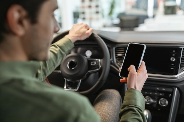 Onherkenbare jongeman die een smartphone gebruikt die in de bestuurdersstoel zit van een nieuwe auto die een voertuig koopt bij auto