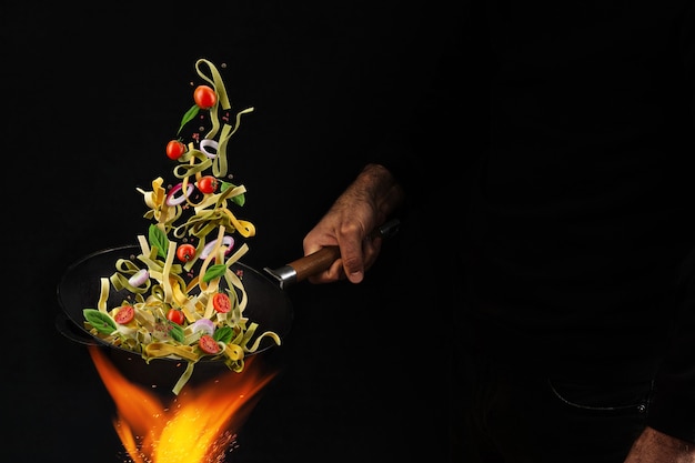 Onherkenbare man die wokpan boven vuur houdt en pasta kookt met cherrytomaatjes, ui en basilicum...