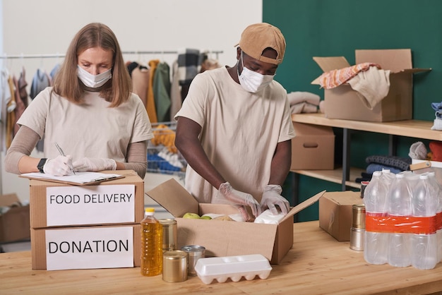 Onherkenbare man en vrouw met maskers op gezichten die aan tafel voedselvoorraden inpakken voor donatie