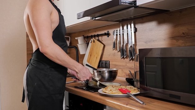 Onherkenbare man met een naakte torso en in een zwart schort bereidt pannenkoeken in de keuken.