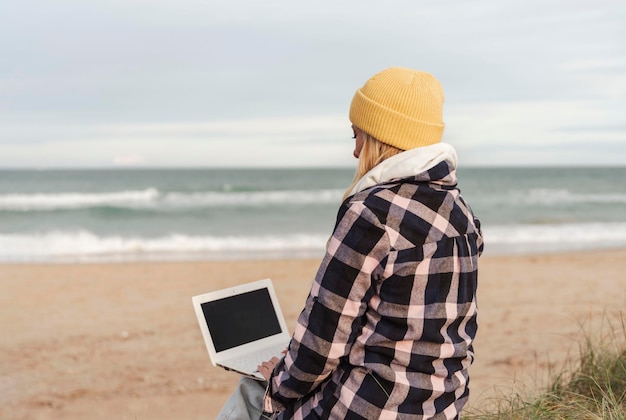 Onherkenbare nomade digitale vrouw in de winter of herfst zittend op het strand met behulp van laptop