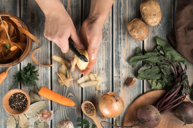 Onherkenbare vrouw die aardappelen schilt met een zwart mes in de buurt van een houten kom met wortelschillen, een kommetje met peperkorrels