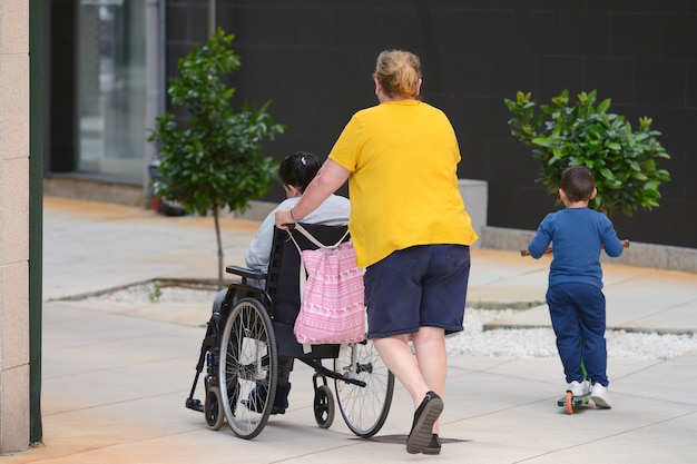 Onherkenbare vrouw die een rolstoel duwt met een gehandicapte persoon, naast een kleine jongen die op een scooter rijdt, achteraanzicht op een stadsstraat