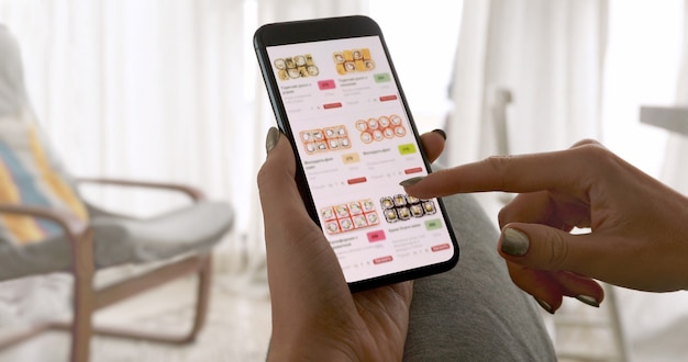 Onherkenbare vrouw met telefoon met app levering sushi eten op scherm thuis