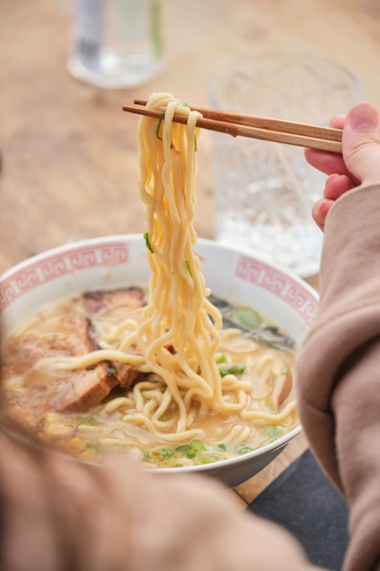 Onherkenbare vrouw zit aan een houten tafel en eet smakelijke ramen soep met noedels en gesneden varkensvlees met stokjes in een Aziatisch restaurant