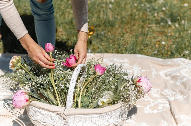 Onherkenbare vrouwenhanden die roze tulpen en wilde witte bloemen aanraken en schikken in een mand op een uitgespreide deken op het gras.