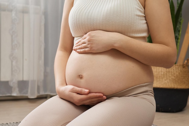 Onherkenbare zwangere vrouw in beige kleding zit op de vloer in de woonkamer en wacht op haar toekomstige baby met liefde en zorgt voor haar toekomstige kind