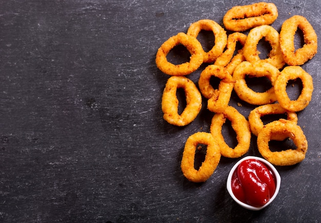 Onion rings with ketchup on dark background, top view