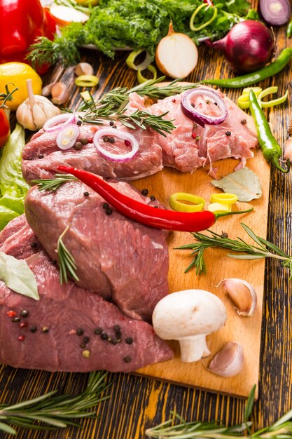 Cipolle, peperoni e funghi su pezzi di carne rossa cruda sul tagliere di legno accanto a rosmarino, lattuga e altre verdure