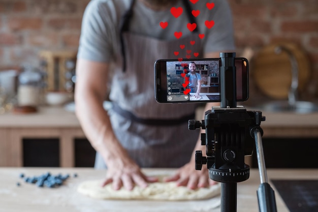 Online baking class Successful culinary vlog Homemade pastry Male chef blogger kneading dough recording video live streaming on smartphone on tripod in blur kitchen with like hearts