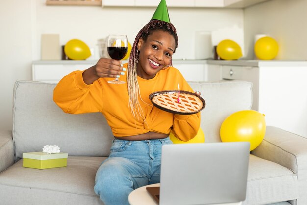 Online birthday Excited african american woman celebrating bday showing pie and glass of wine video calling on laptop