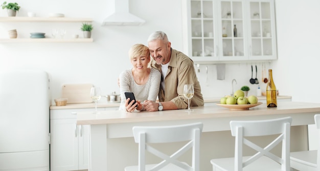 Online chat, call and communication with family remotely and celebrate at home during covid-19 quarantine. Smiling adult couple look at smartphone in kitchen interior with apples and glasses of wine