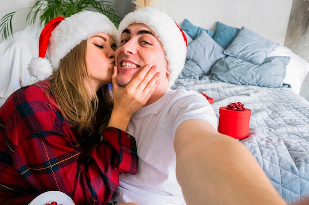 Online Christmas Party. Woman kissing man taking selfie wearing santa hats