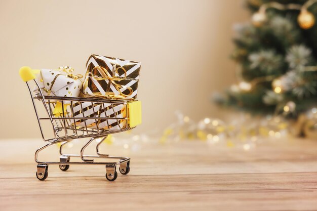 Online christmas shopping and sale Shopping cart with gift boxes tied golden ribbons and bow on wooden background Supermarket trolley full presents Copy space