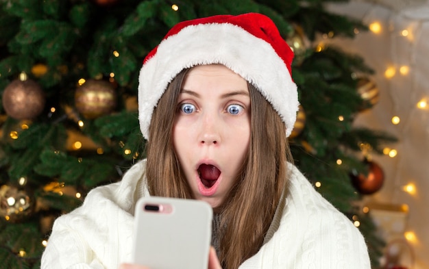 オンラインクリスマスショッピング。サンタの帽子をかぶったショックを受けた少女が携帯電話を持っています。