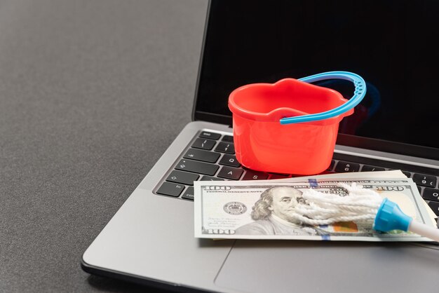 Photo online cleaning service toy bucket mop and cash dollars on laptop keyboard concept of money laundering online