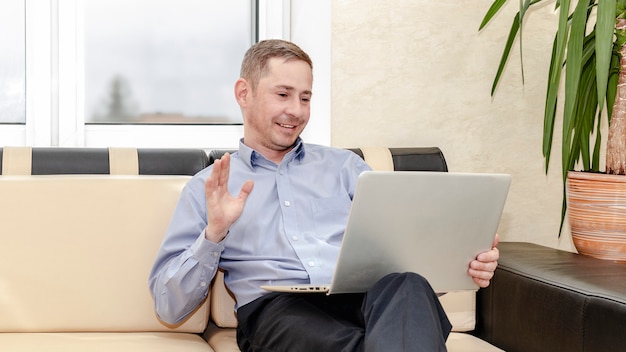 Online communicatie. Een jonge manager in een blauw shirt die op de bank zit, communiceert met een laptop op schoot.