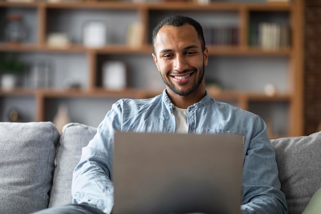 Online communicatie knappe jonge zwarte man zittend op de bank en met behulp van Laptop
