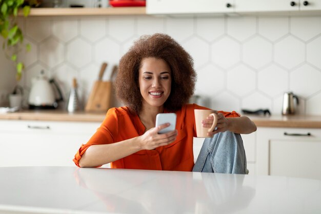 オンラインコミュニケーション美しい黒人女性がスマートフォンを使用し、キッチンでコーヒーを飲む