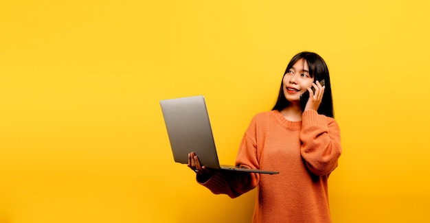 온라인 커뮤니케이션 개념 아시아 소녀는 집에서 노트북을 사용하는 노란색 스튜디오 사진 그녀는 소셜 네트워크, 쇼핑 또는 온라인 작업에서 친구들과 채팅하면서 온라인에서 자유 시간을 보내는 것을 기쁘게 생각합니다.