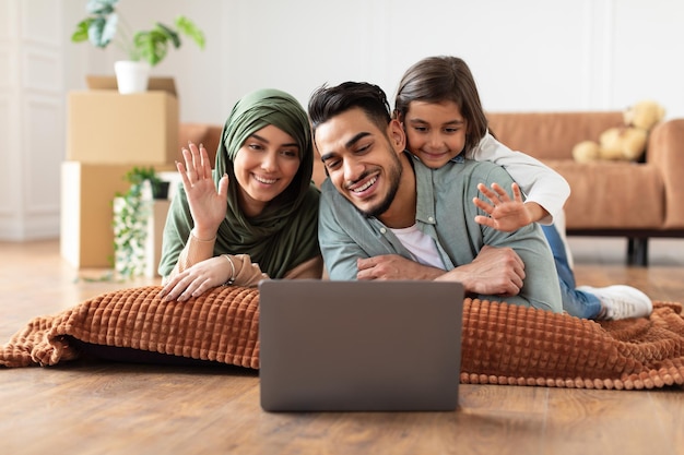 Online communication. smiling arab man, woman in hijab and\
little girl making digital video chat with friends or family using\
laptop, waving to webcam, sitting on the floor carpet in living\
room