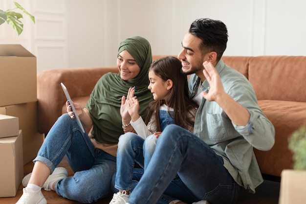 Online Communication. Smiling arab man, woman in hijab and little girl making digital video chat with friends or family using tablet, waving to webcam, sitting on the floor in living room