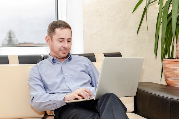 온라인 커뮤니케이션. 파란색 셔츠를 입은 젊은 매니저가 소파에 앉아 무릎에 노트북을 들고 의사소통을 합니다.