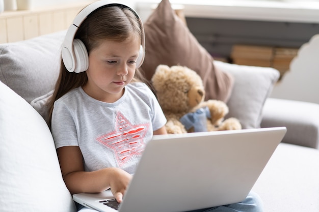 子供のオンライン教育。自宅のソファに座っているビデオレッスン教師会議ラップトップを探しているヘッドフォンを持つ甘い女の子。