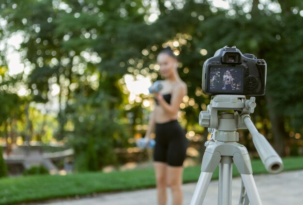 オンラインフィットネストレーナー。若い魅力的なフィットの女性は、朝、屋外のカメラでブログを撮影し、ダンベルで運動する方法を示しています。スポーツブログ、フィットネスvlog