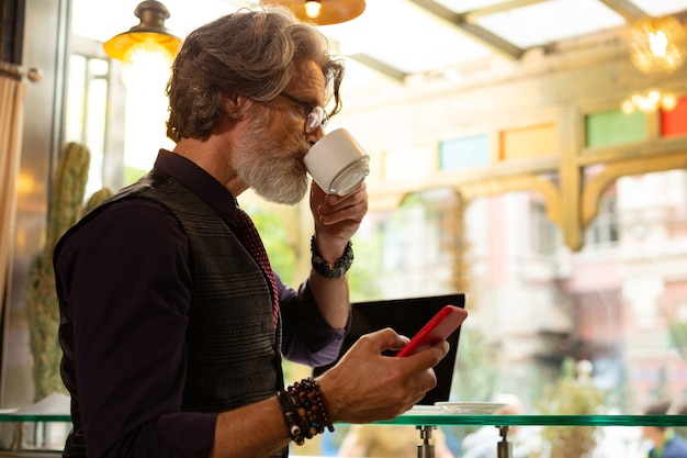 Online gesprekken. Ernstige man die koffie drinkt in het café en nadenkt over het reageren op een bericht op zijn smartphone.