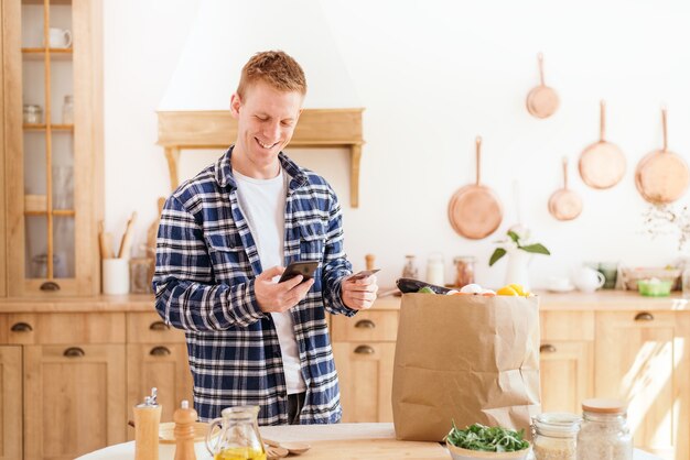 Online Grocery Shopping A male using a credit card and a phone pays for food products standing in the kitchen at home