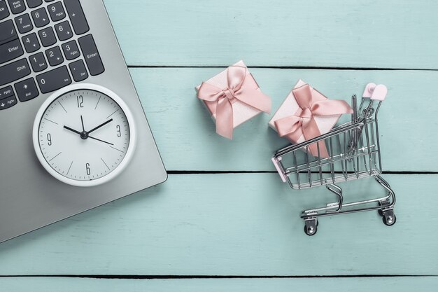 Online, holiday shopping. Supermarket trolley with gift boxes, laptop and clock on blue wooden surface. Top view