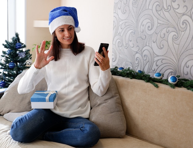 Online kerstgroeten. Een vrouw met een telefoon thuis in een kerstmuts feliciteert vrienden.