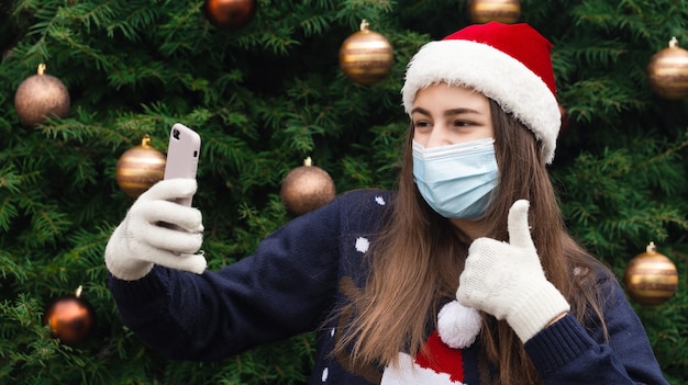 Online kerstgroeten. sluit omhoog portret van vrouw die een hoed van de kerstman en medisch masker met emotie draagt. tegen de achtergrond van een kerstboom. coronapandemie