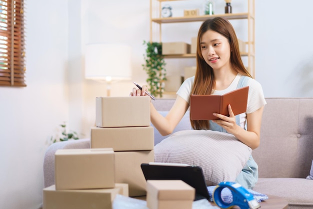 Online merchant concept Female entrepreneur writes on parcel box after reading address on notebook