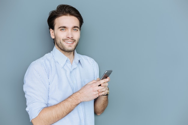 Online messaging. Handsome positive young man looking at you and smiling while typing a message on his smartphone