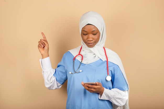 Online nurse with headscarf isolated on yellow background.Holds smartphone, points finger up