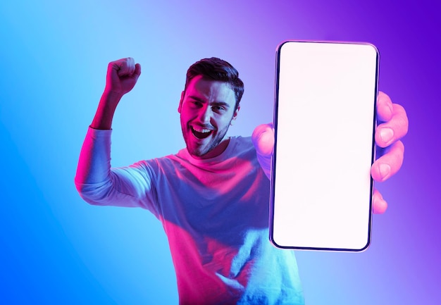 Online offer excited young man showing blank smartphone and celebrating success