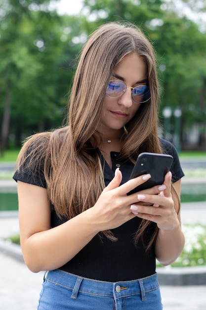 Online onderwijs, freelance, zaken, technologie en internetconcept - Een Europees donkerharig meisje in vrijetijdskleding staat in een park in de zomer, houdt haar telefoon vast en kijkt erin.